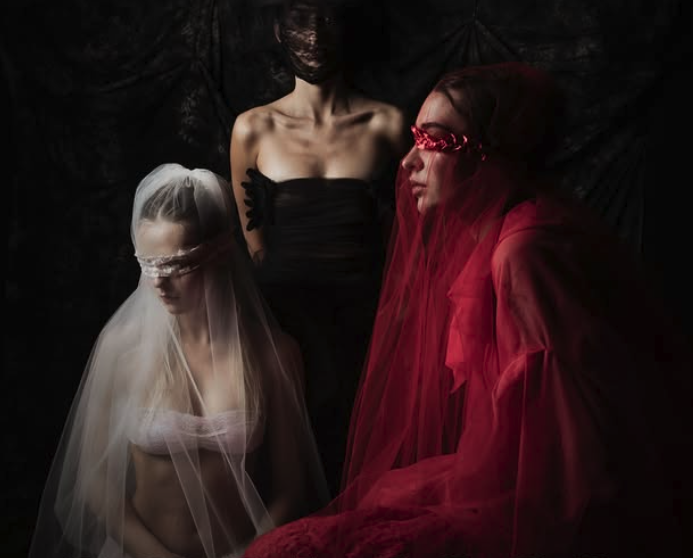 A dark, moody artistic portrait of three women whose eyes and faces are obscured by sheer white, black, and red veils