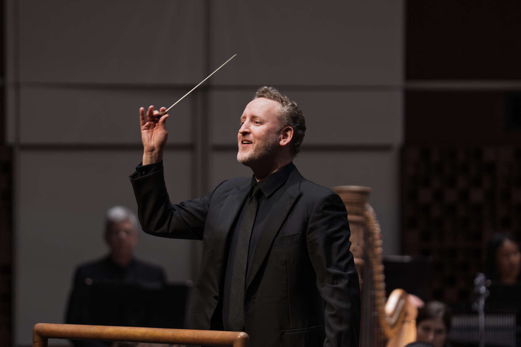 Michael Francis, Music Director of The Florida Orchestra, in a side-profile portrait. The person is smiling and looking upward, wearing a professional black suit, black dress shirt, and a black tie against a neutral grey background.