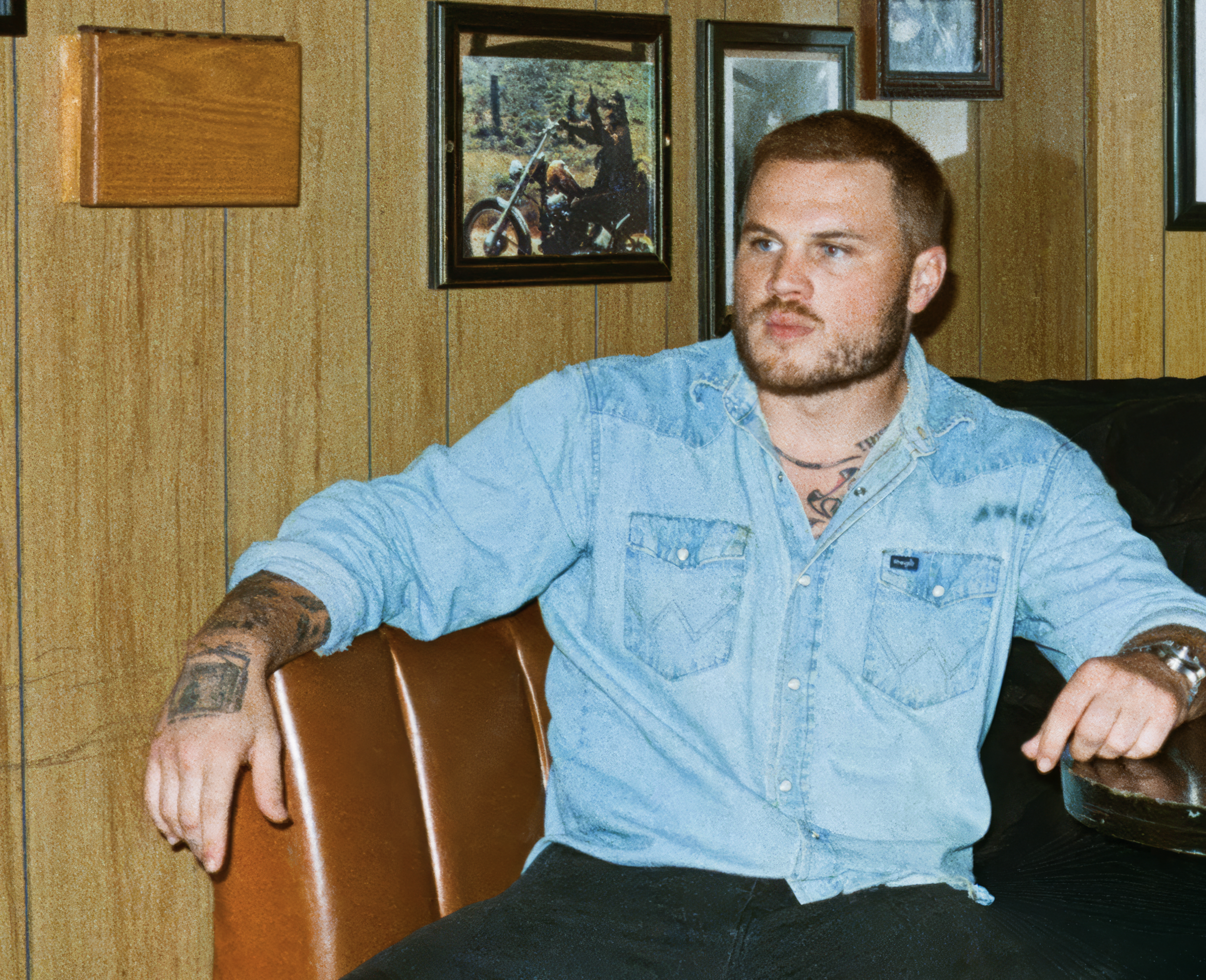 Musician Zach Bryan wearing a light blue denim shirt and sitting on a brown leather couch in a room with wood-paneled walls. The person has visible tattoos on their arms and looks off-camera; framed photos are on the wall behind them.
